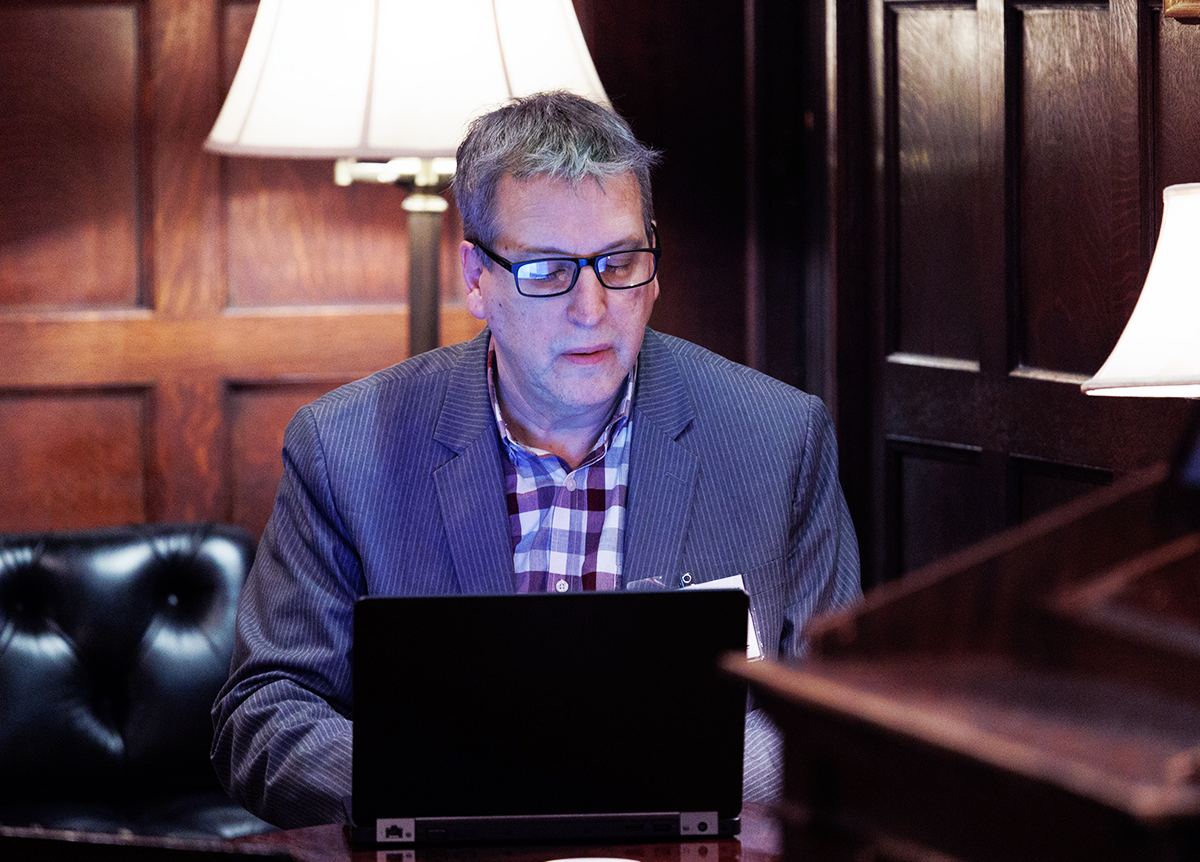 A BISG member sitting at a table in a dark wood office space, looking at a laptop screen, his face lit up by the glow. 