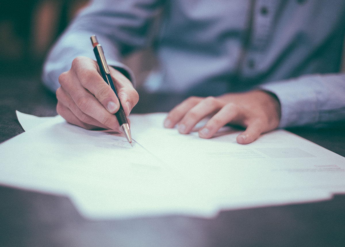 Closeup of hands writing on a pile of papers. Camera is focused on the right hand, holding a pen.