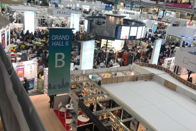 A view of a covention space from a second-flooor balcony. Crowds are gathered around booths, and banners hang from the ceiling. 