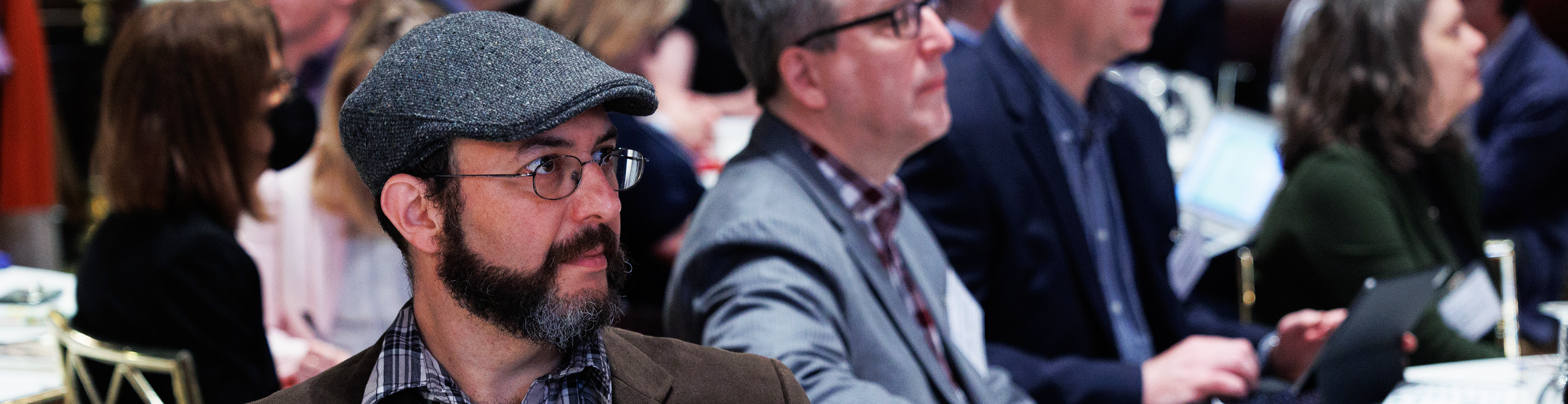 A table at a BISG event, the camera is focused on a single man intently looking off-camera at a speaker.