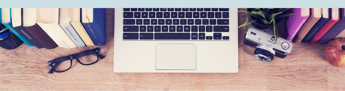 View from above of a desk with an open laptop, books lined up neatly, a pair of glasses, a camera, and apple, and a small pottted plant. The laptop is cropped at the keyboard, and most of the space is empty desk. 