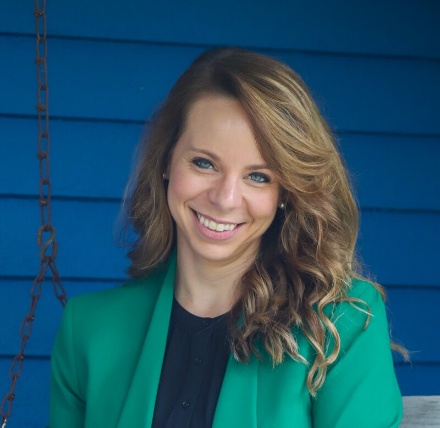 A woman with long, wavy hair and a green jacket is standing against a blue wall.AI-generated content may be incorrect.