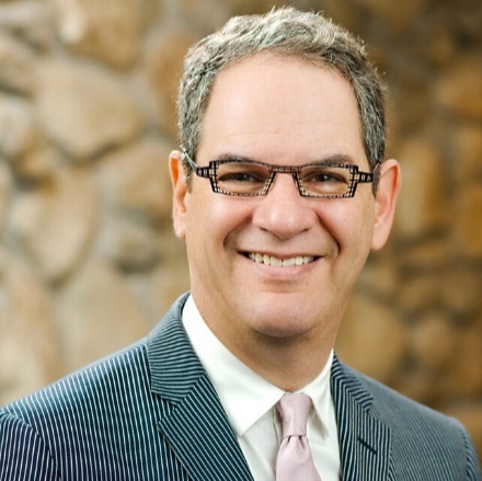 The image shows a person dressed in a striped suit and a pink tie, standing in front of a textured stone wall.AI-generated content may be incorrect.