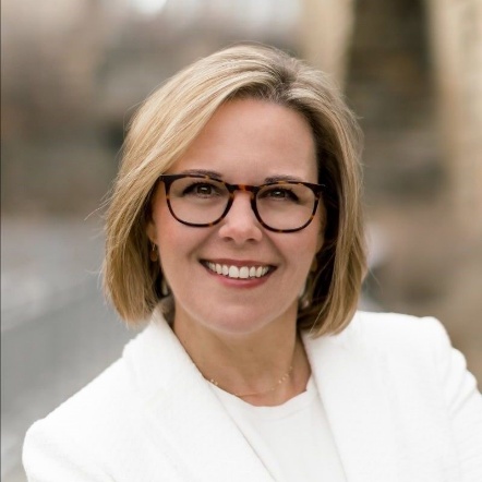 The image shows a person with short, light-colored hair, wearing a white, high-collar shirt, and a black jacket.AI-generated content may be incorrect.