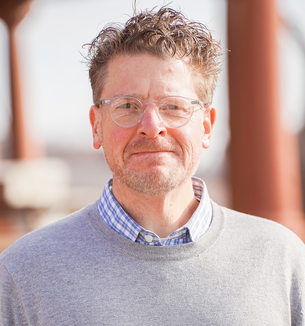 Headshot of program presenter, Joe Benesh, standing outside with a blurred background, wearing a gray sweater over a checkered collared shirt