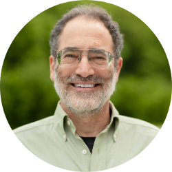 Headshot of presenter, Andy Robinson, wearing glasses and a green collared shirt, standing in front of a blurred outdoor background