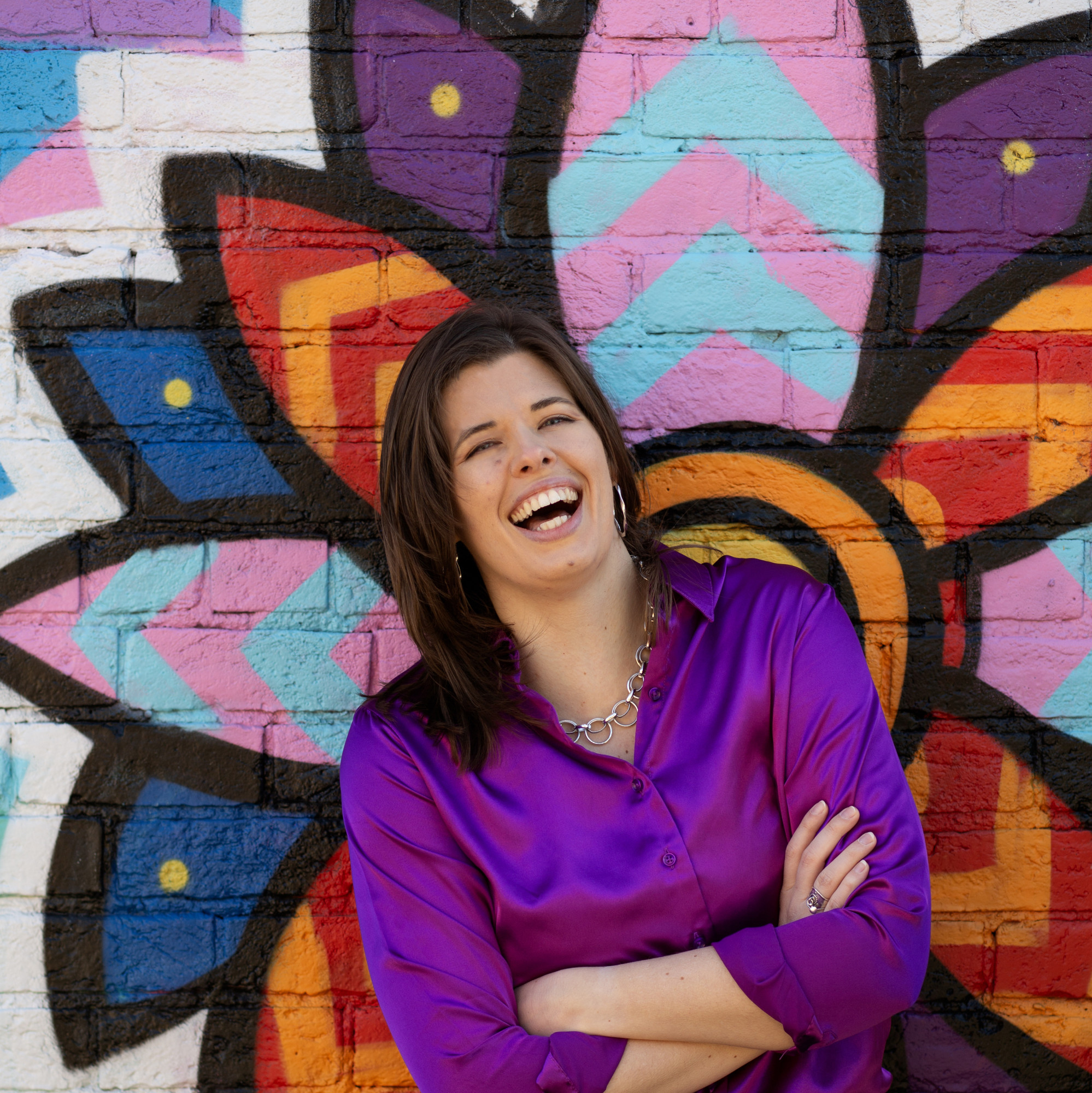 Headshot of program presenter, Chelsea Frost, laughing, wearing a purple top, standing in front of a multi-color flower mural
