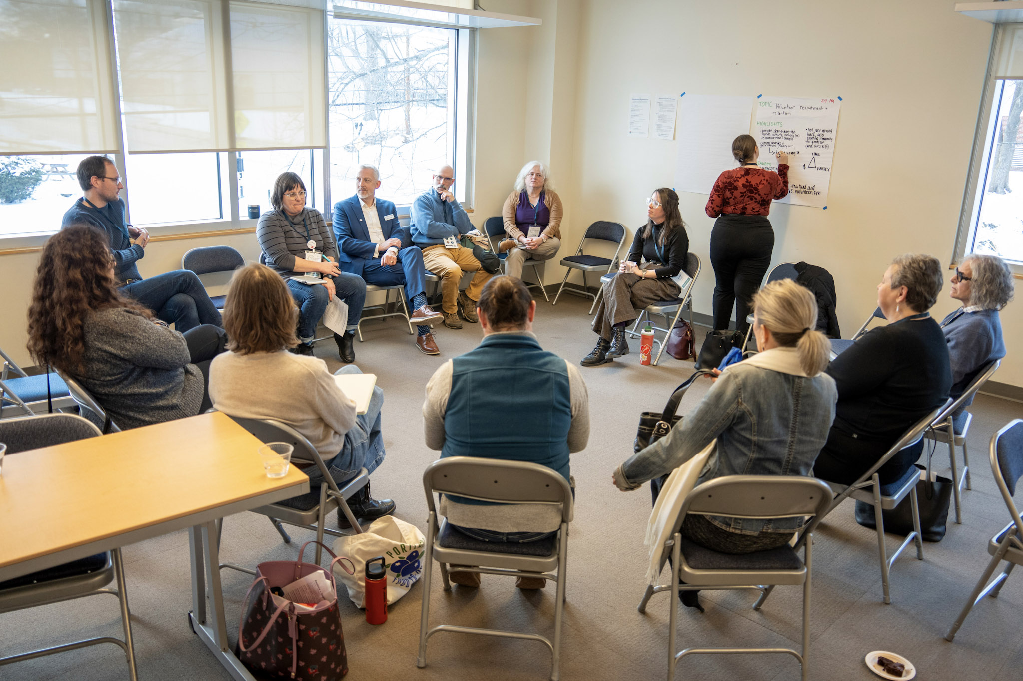 A seated circle of event participants engaged in conversation, while one person takes notes on a flip chart on the back wall.
