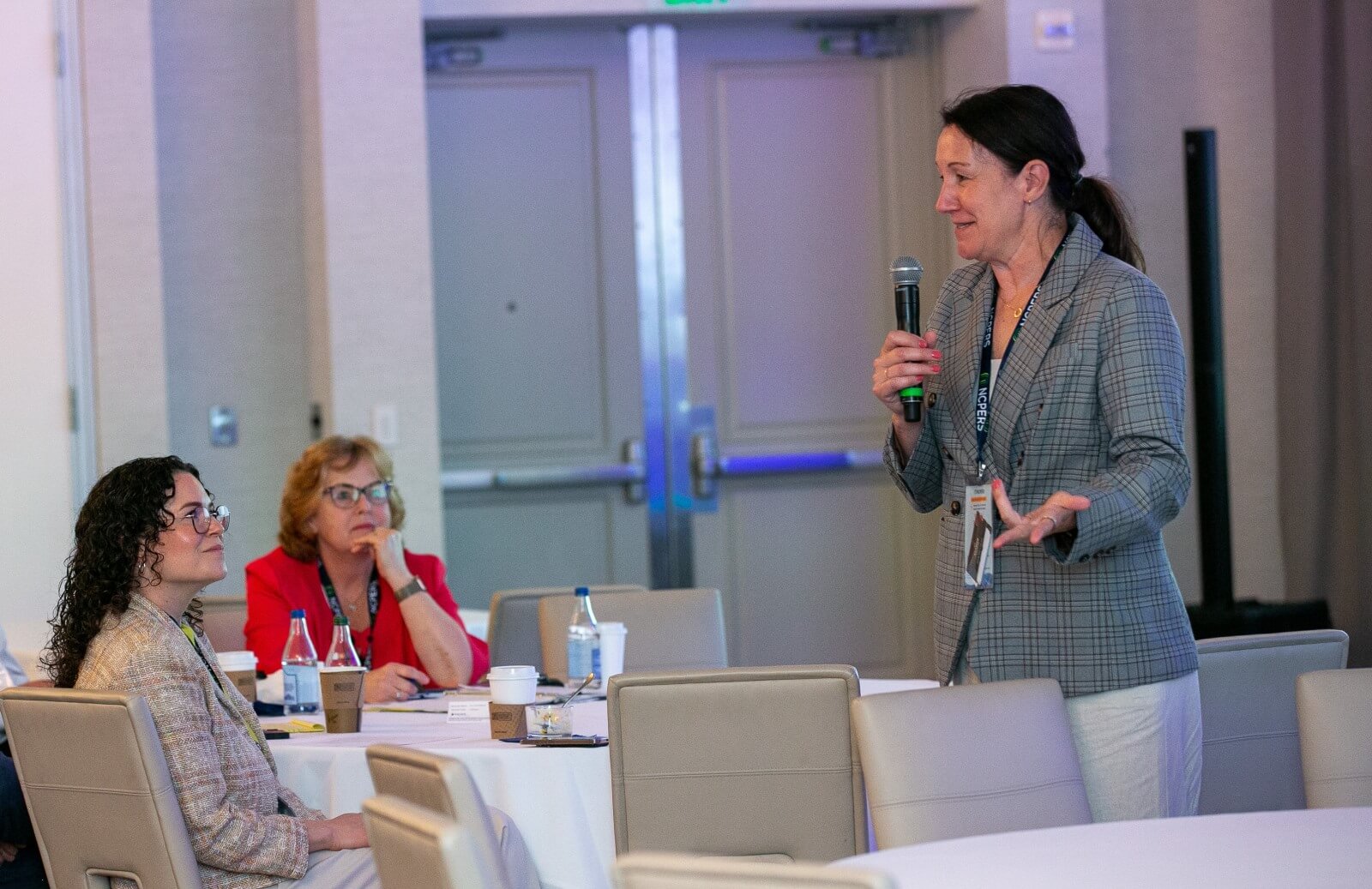 A presenter holding a microphone close to a roundtable with two seated attendees