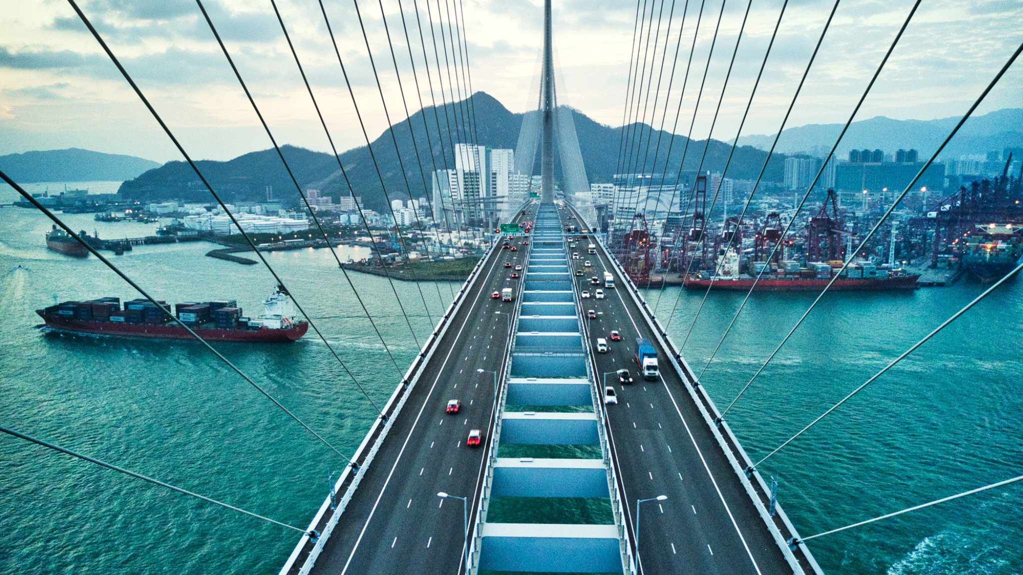Aerial view of a cable-stayed bridge over water leading toward a port and mountains.