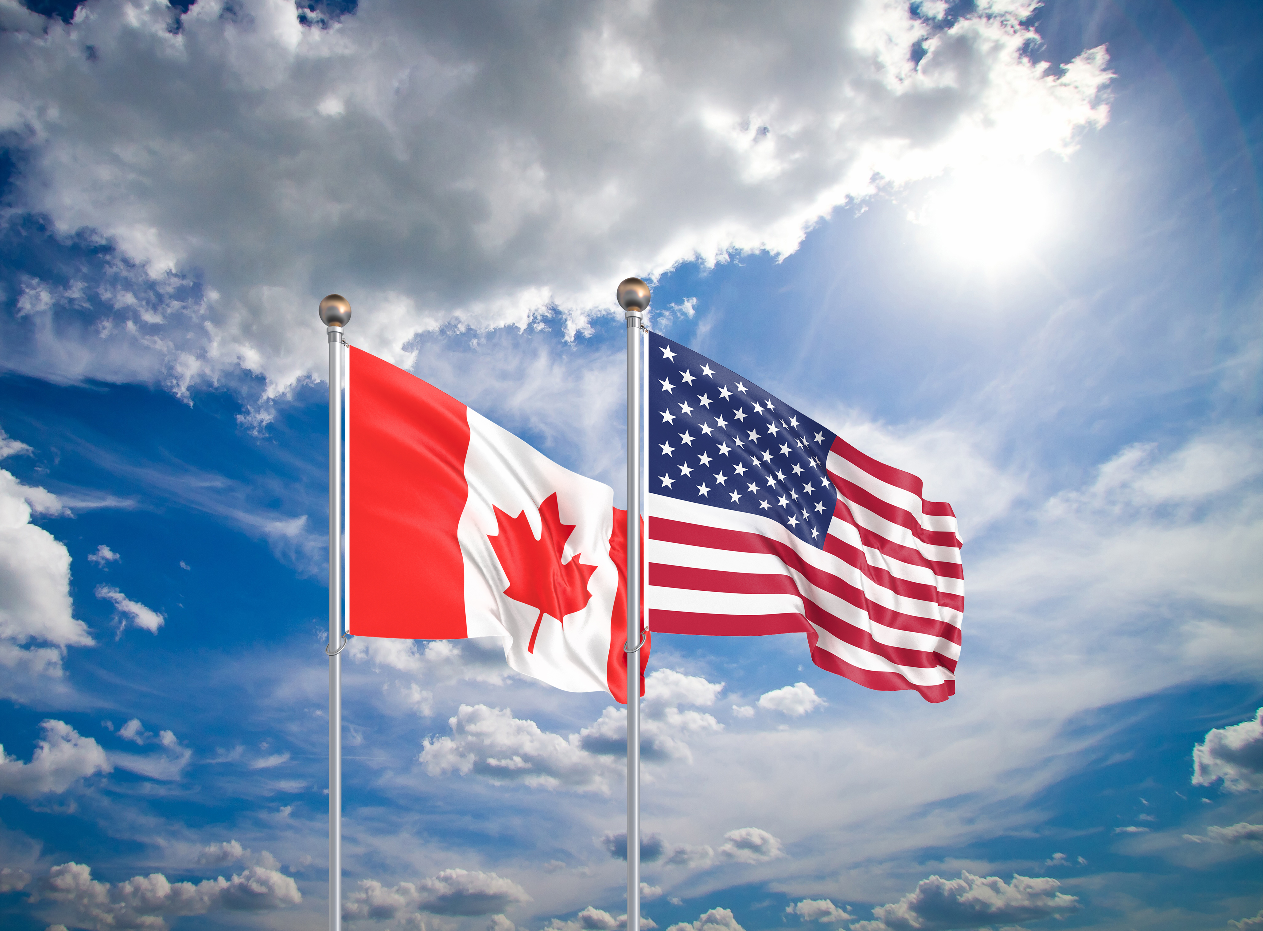 Canadian and American flags waving side by side against a blue sky with clouds