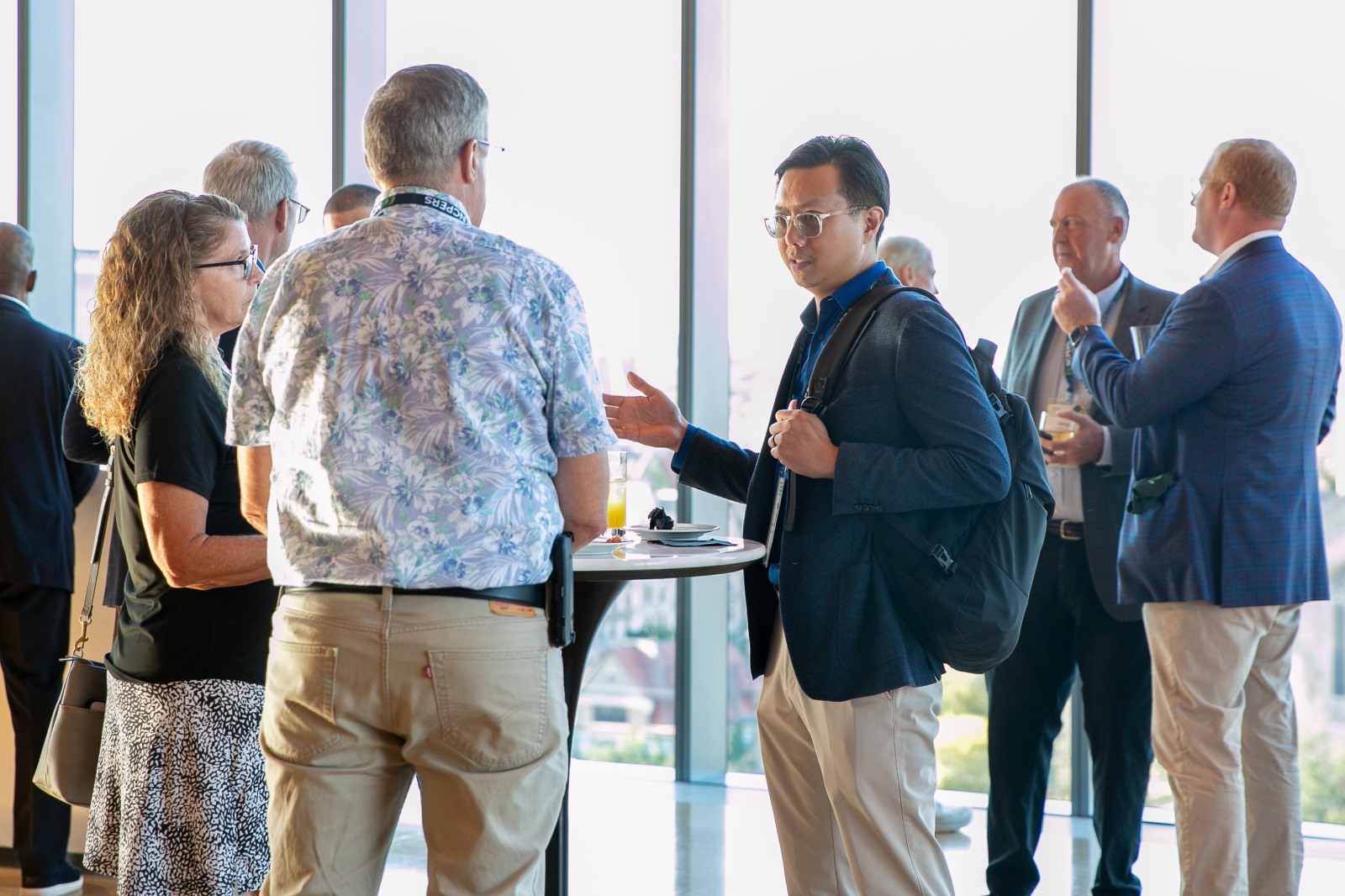 Attendees standing in conversation around a high-top table in a sunlit lobby with floor-to-ceiling windows overlooking the city