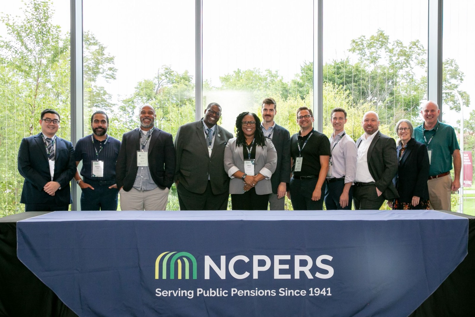 Eleven competition participants, mentors, and judges stand smiling behind an NCPERS table at the Pension Lab: NextGen Competition.
