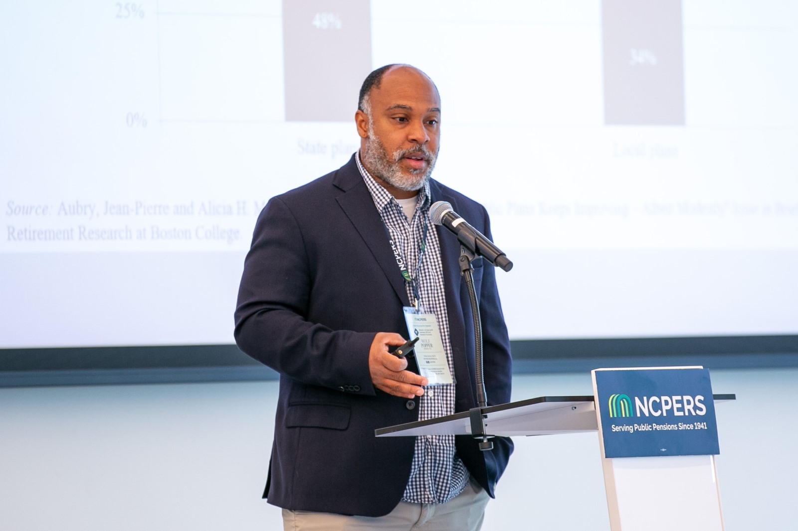 A speaker stands at an NCPERS podium with a microphone, presenting in front of a projected bar chart comparing state and local pension plans