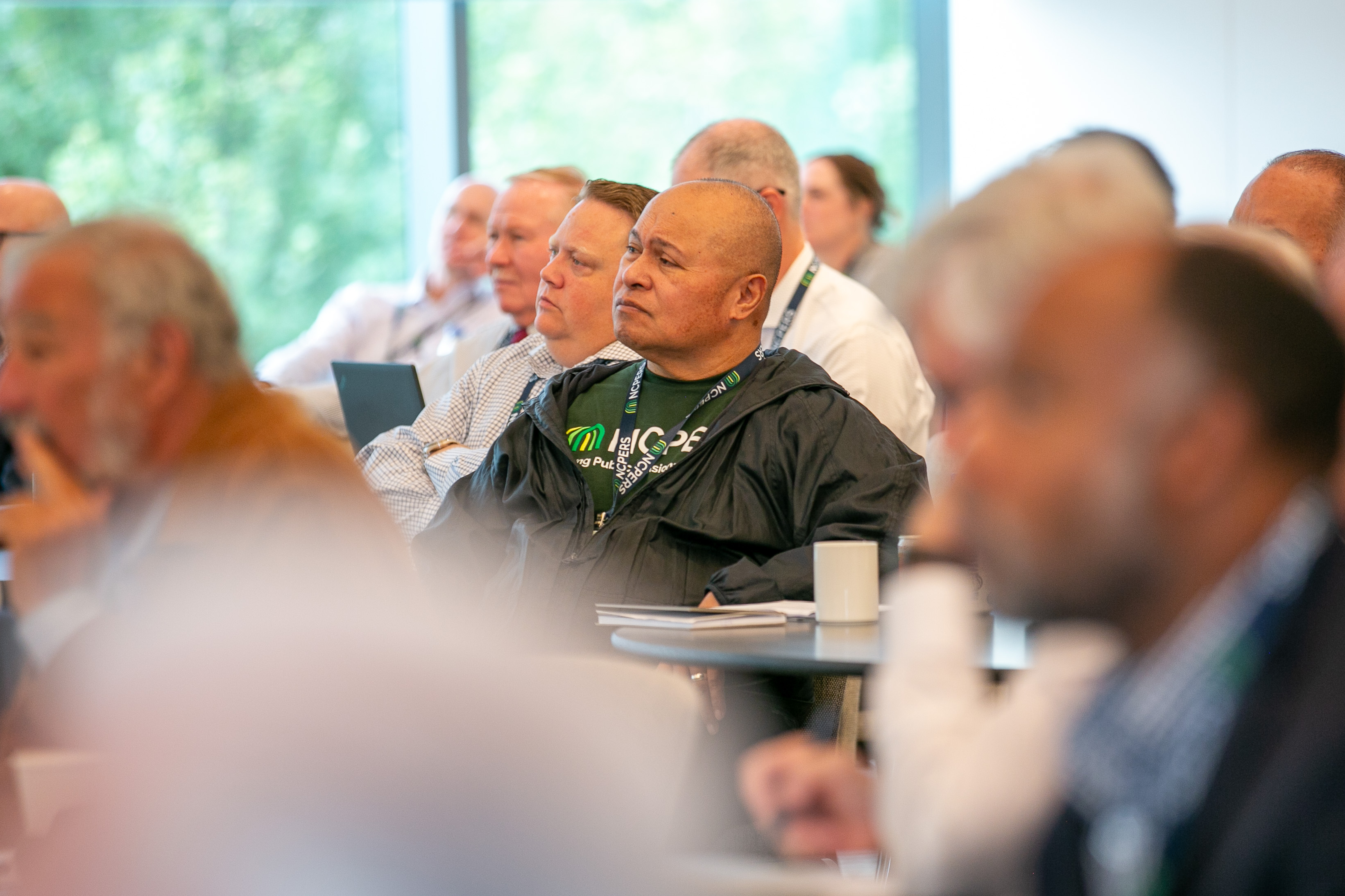 Attendees seated at round tables in a session room, turned toward a presenter off-camera; one wears an NCPERS shirt and conference lanyard