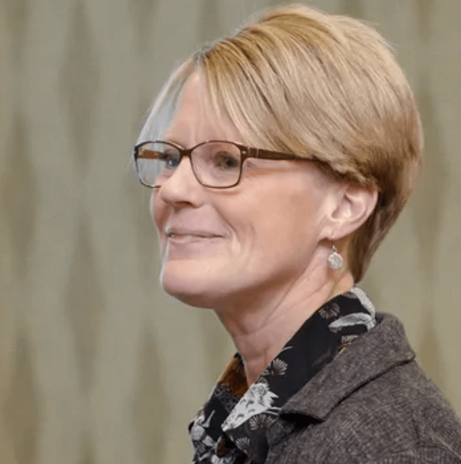 Photo of Kim Aycock, a blond woman with short hair wearing brown glasses, a black scarf, and brown collared shirt who is smiling and looking off to the left of the frame