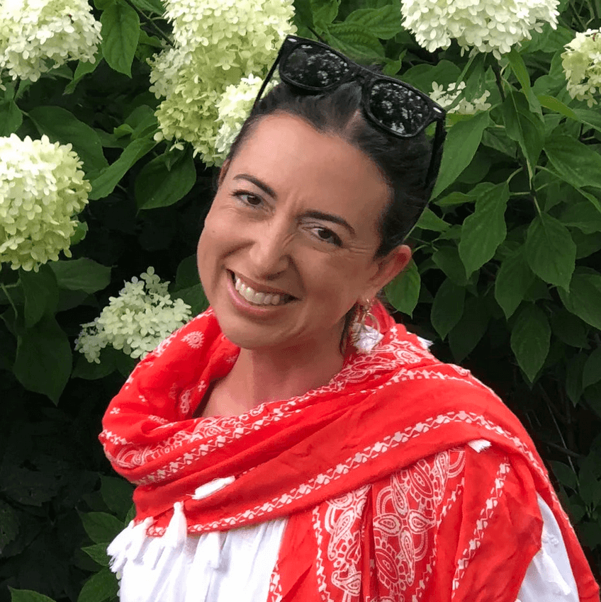 Photo of Jolly Corley, a woman with brown curly hair that is tied up in a bun, smiling at the camera with greenery behind her. She is wearing a red scarf and white shirt.